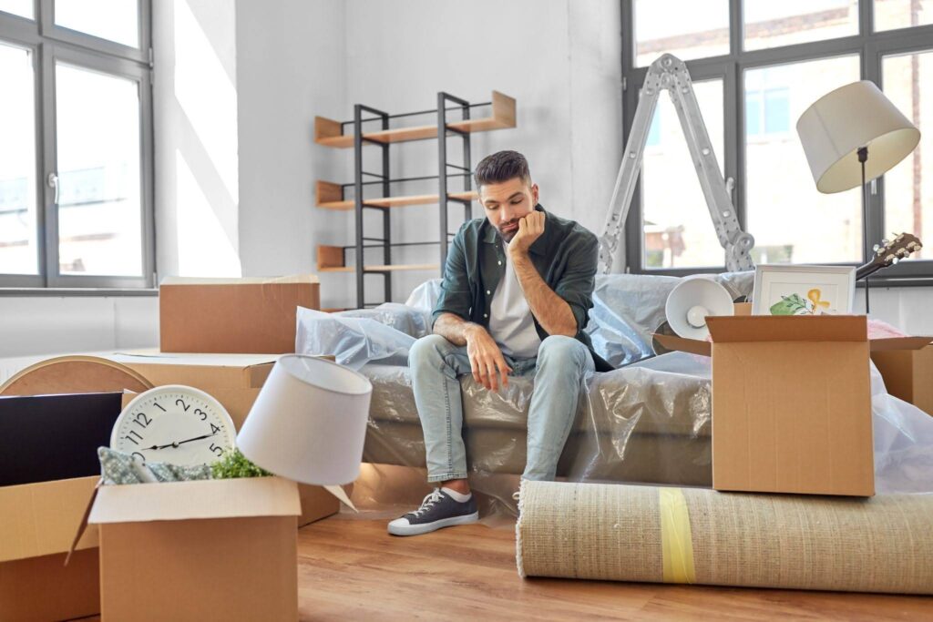 a sad man sitting in the middle of the room surrounded by cardboard boxes for moving